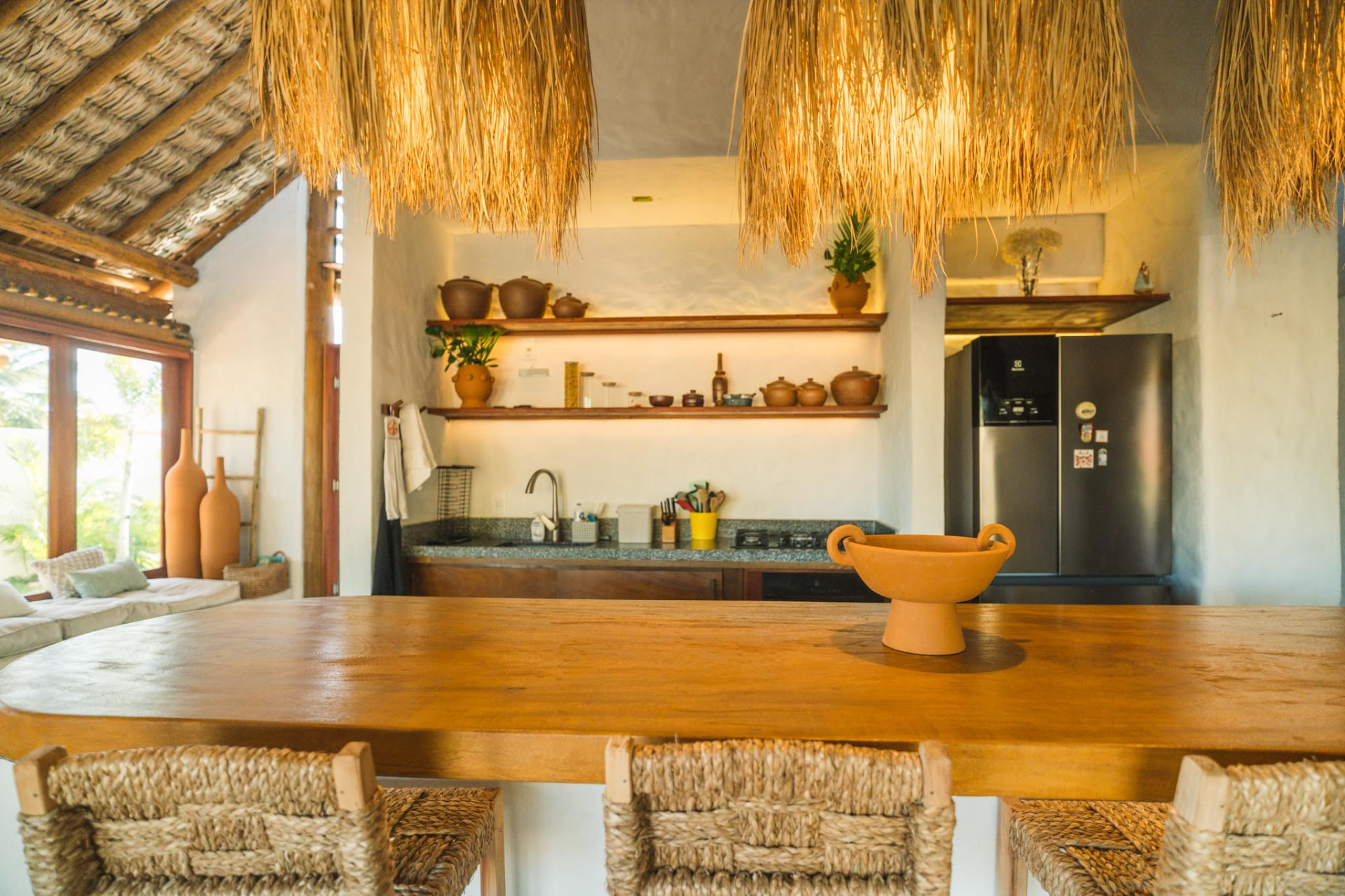 Cozinha integrada com mesa de madeira, cerâmicas e cobertura de palha